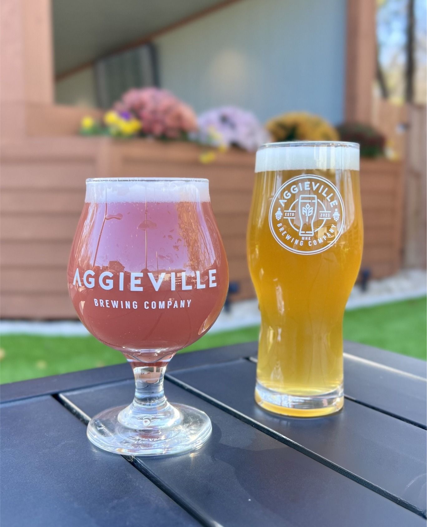 Two glasses of beer, one pink and round, the other tall and golden, set on a table in a vibrant garden atmosphere.