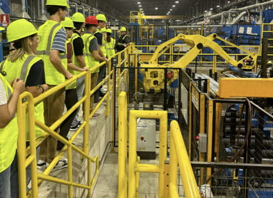 A group in safety gear tours a manufacturing facility, evoking curiosity, safety, teamwork, and professionalism.