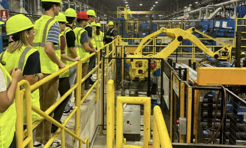 A group in safety gear tours a manufacturing facility, evoking curiosity, safety, teamwork, and professionalism.