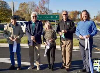 Five people in smart-casual attire prepare to cut ribbons at an outdoor event, symbolizing celebration and community pride.