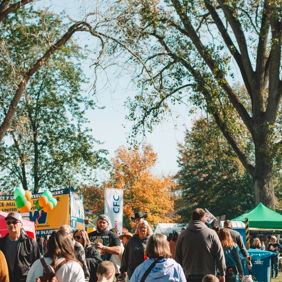 Lively outdoor community event with families walking along a tree-lined path, colorful booths, and a cheerful atmosphere.