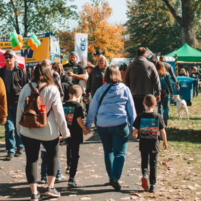 Lively outdoor community event with families walking along a tree-lined path, colorful booths, and a cheerful atmosphere.