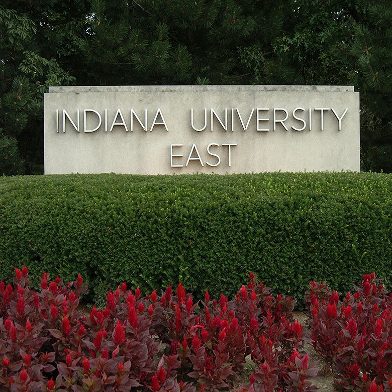 Stone sign "INDIANA UNIVERSITY EAST" amid lush greenery and vibrant plants, conveying a welcoming and tranquil campus atmosphere.