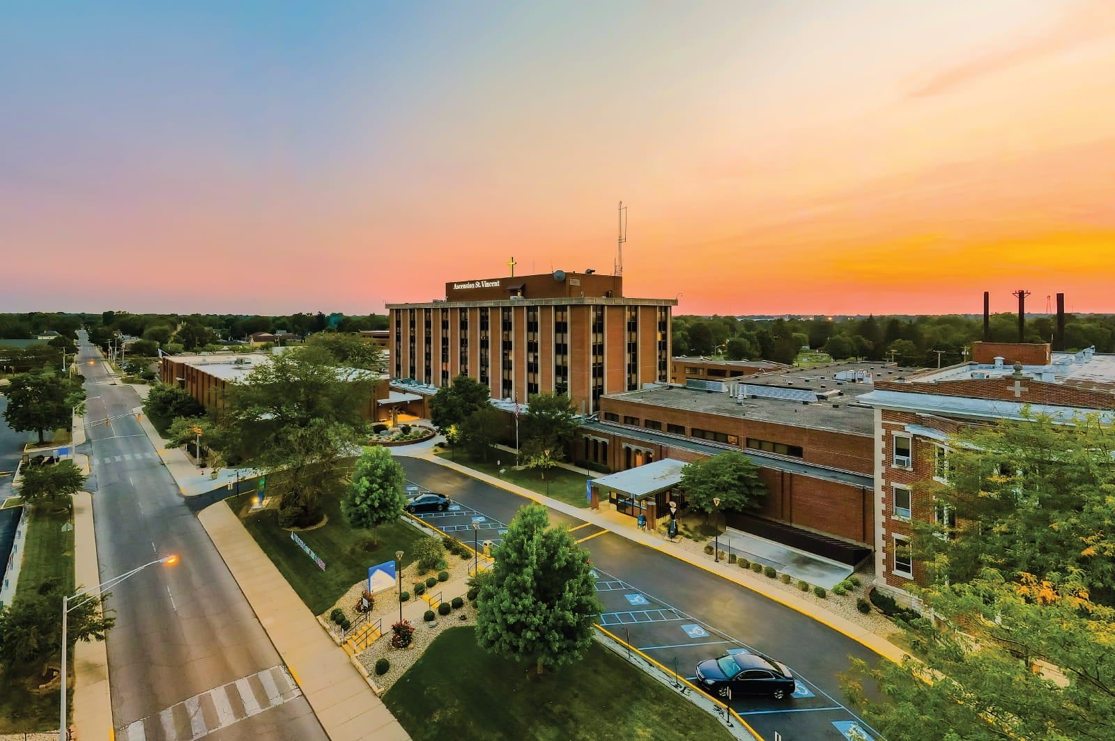 Sunset view of a hospital with vibrant skies, trees, and parked cars, evoking calmness, hope, and a sense of care.
