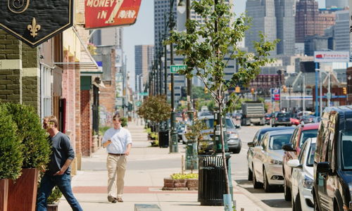 Bustling urban scene with shops, pedestrians, and modern skyline; evokes liveliness and community on a sunny day.