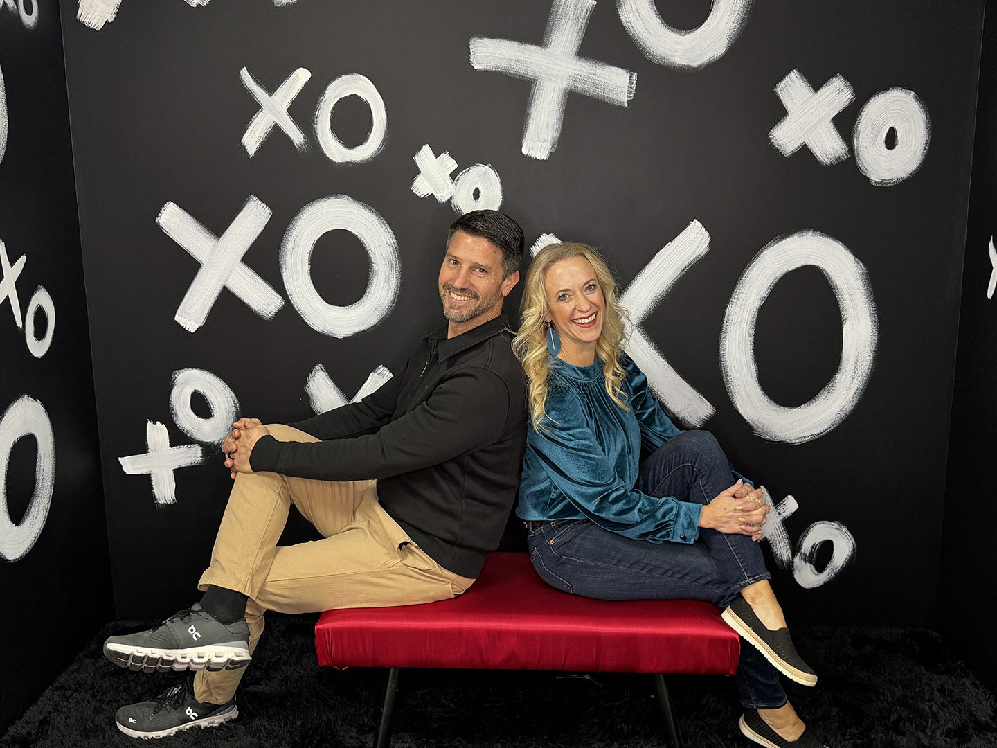 Two individuals sit playfully on a red bench in front of a black wall with "XO" symbols, conveying warmth and joy.