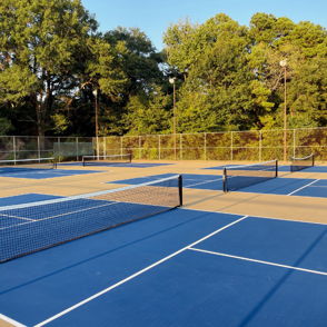 Tennis courts with blue surfaces and black nets, surrounded by green trees, evoking tranquility and a welcoming atmosphere.