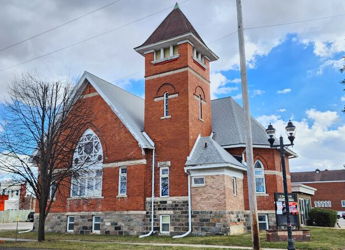 Traditional brick church with steeple, arched window, greenery, and a partly cloudy sky, evoking serenity and community.
