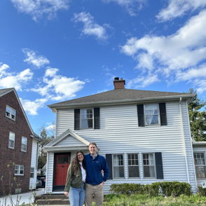 Two smiling individuals stand in front of a white house with a red door, evoking joy and warmth on a sunny day.