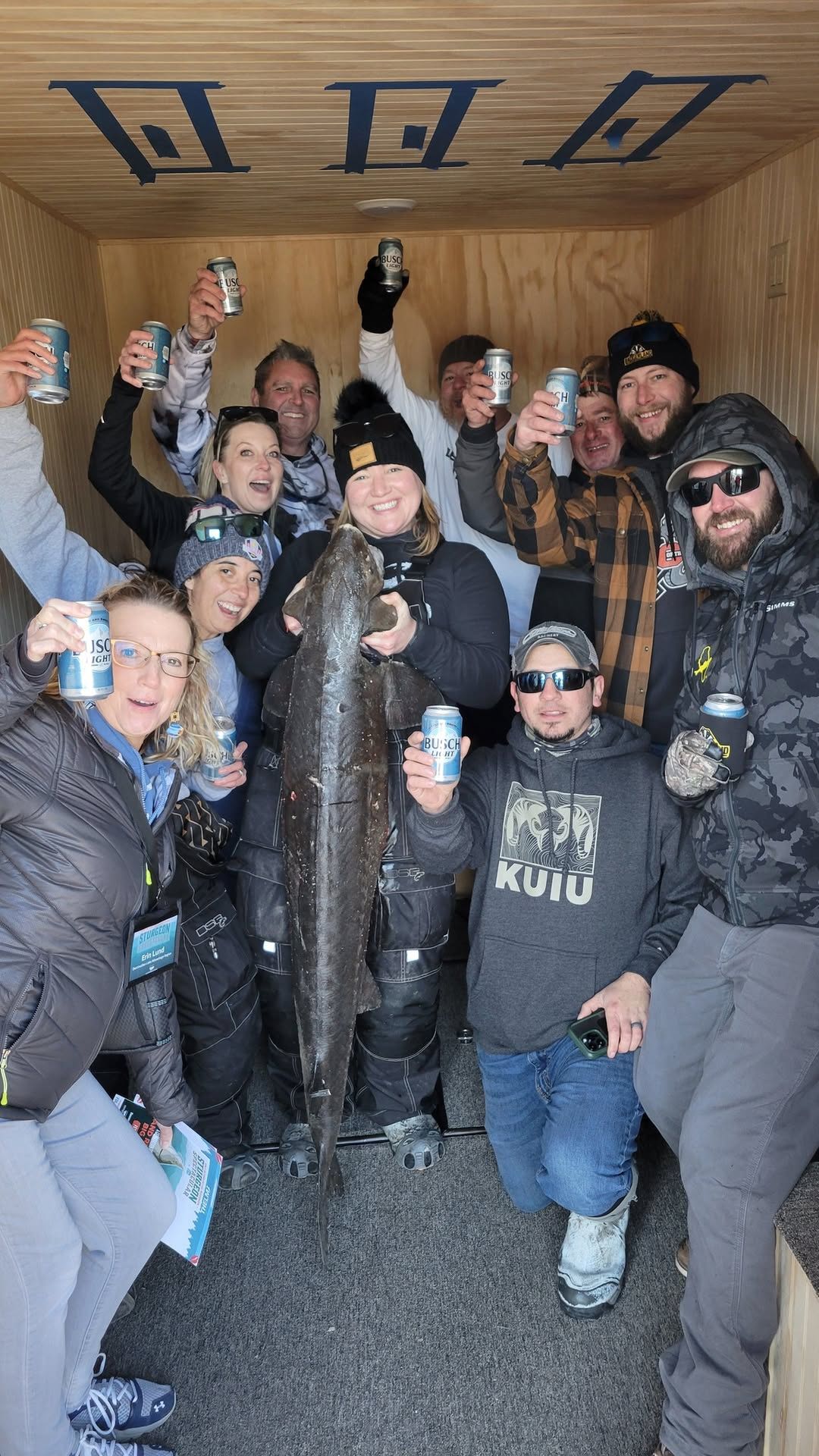 Group of people celebrating indoors with drinks; one holds a large fish, capturing a festive, joyful fishing trip moment.