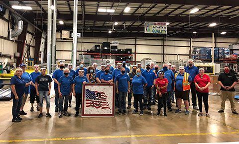 Group of people in blue shirts celebrating, holding an American flag quilt, evoking unity, pride, and teamwork.