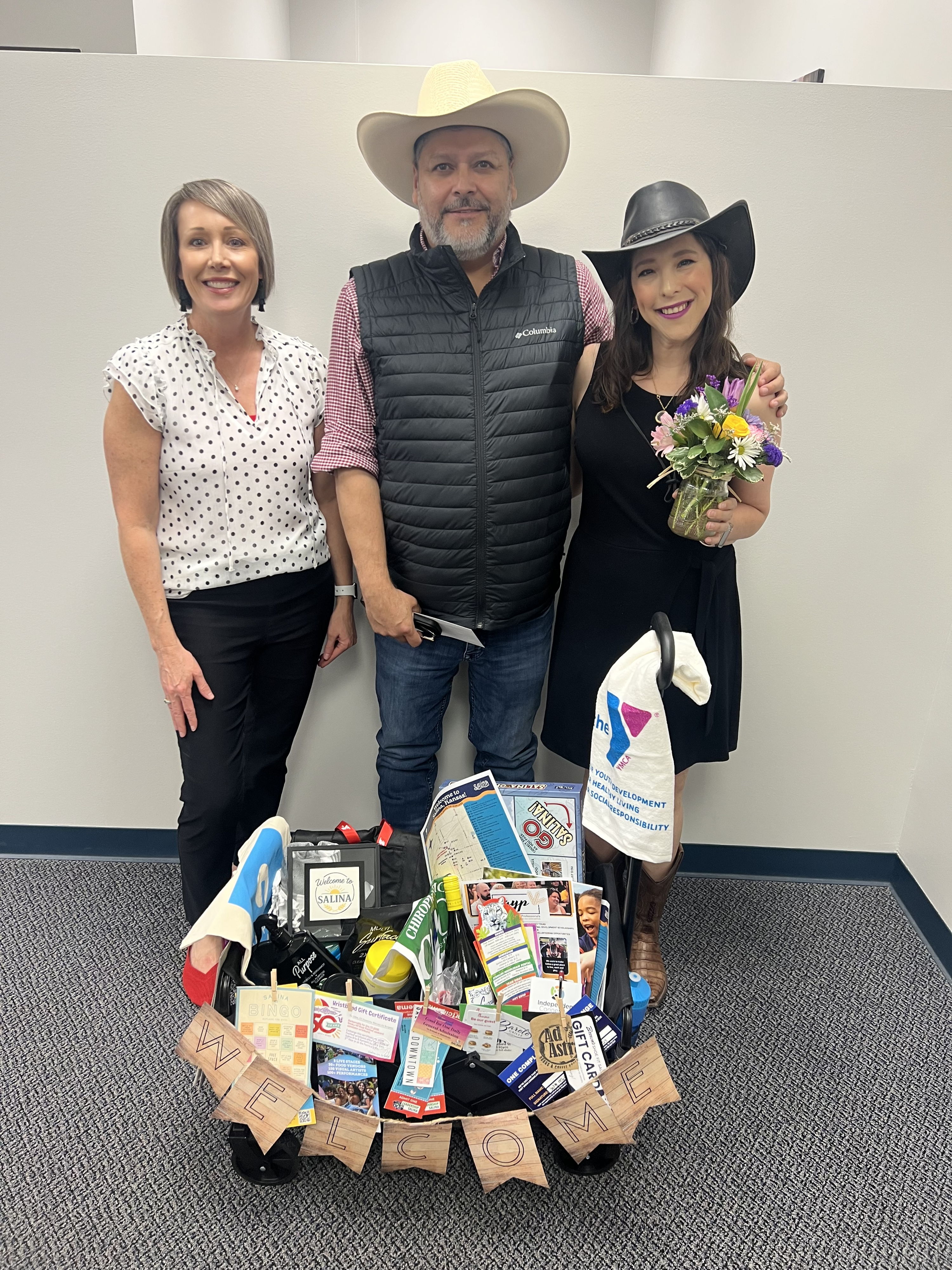 Three smiling people pose together, surrounded by personal items and gifts, capturing a joyful and cheerful celebration.