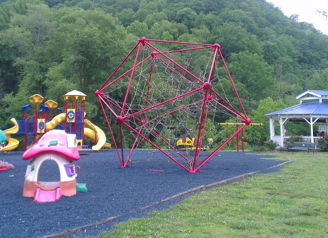Playground with a colorful climbing dome and house-shaped slide, set in vibrant green hills, evokes joy and childhood nostalgia.