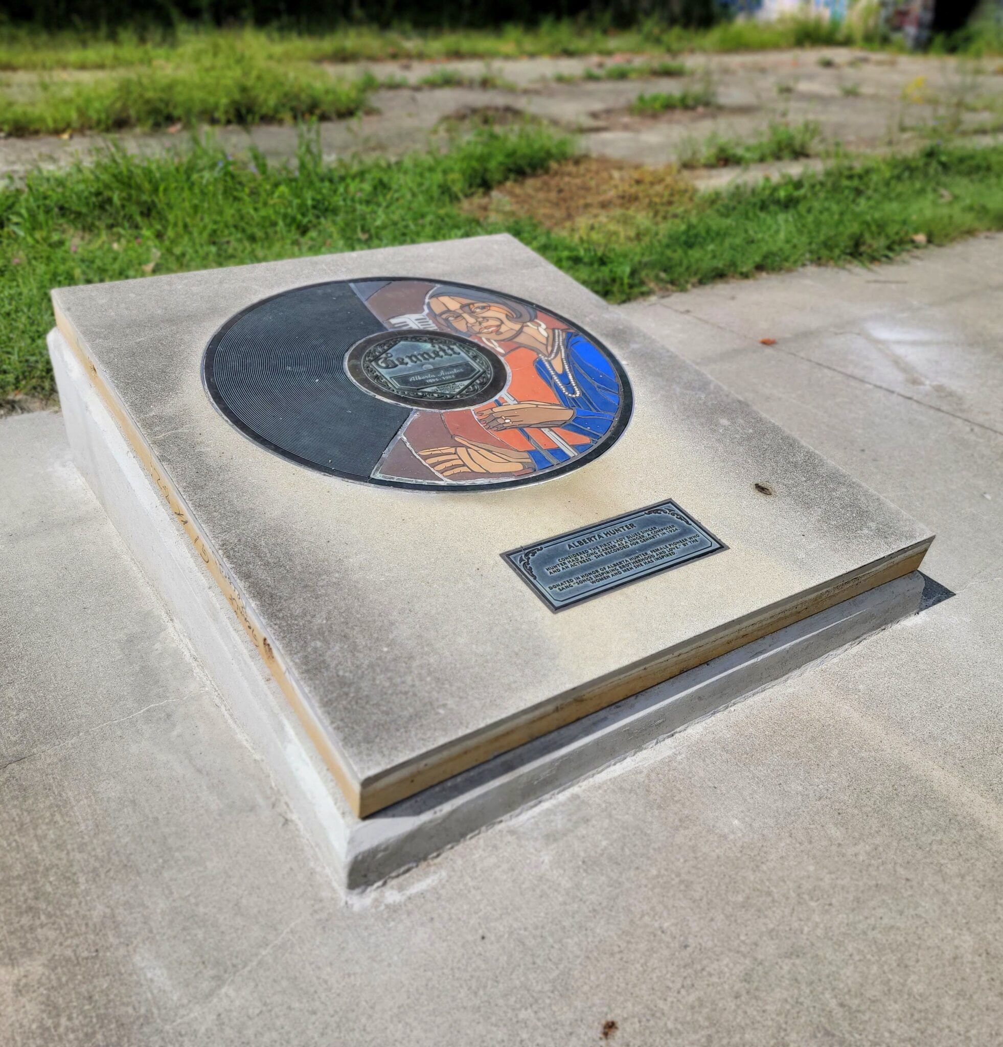 A circular vinyl-record-like plaque honors an individual, set in overgrown surroundings, evoking nostalgia for forgotten history.