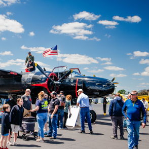 A vintage aircraft in black and red attracts a diverse crowd at a festive outdoor aviation event, evoking excitement and nostalgia.