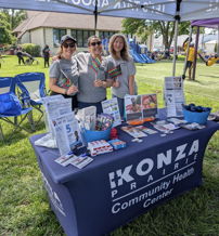 Community health center booth outdoors with smiling women, colorful materials, and a friendly atmosphere, inviting community engagement.