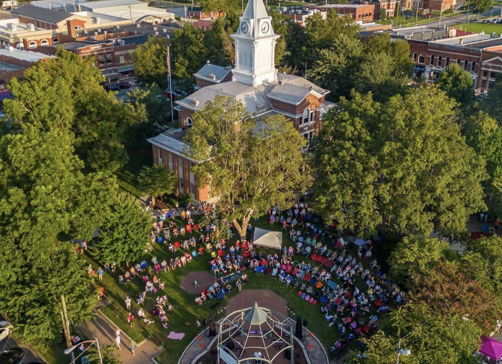 Aerial view of a park with a gazebo and crowd, featuring a town's clock tower, vibrant greenery, and a festive atmosphere.