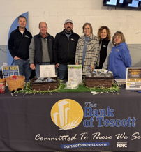 Group of six friendly individuals at a Bank of Tescott table, promoting community engagement with smiles and branded materials.