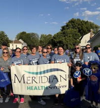 A joyful group in matching gray shirts holds a "Meridian Health Services" banner, celebrating community unity outdoors.