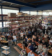 A large group attentively listens to a speaker in a well-lit warehouse, fostering engagement and collaboration.
