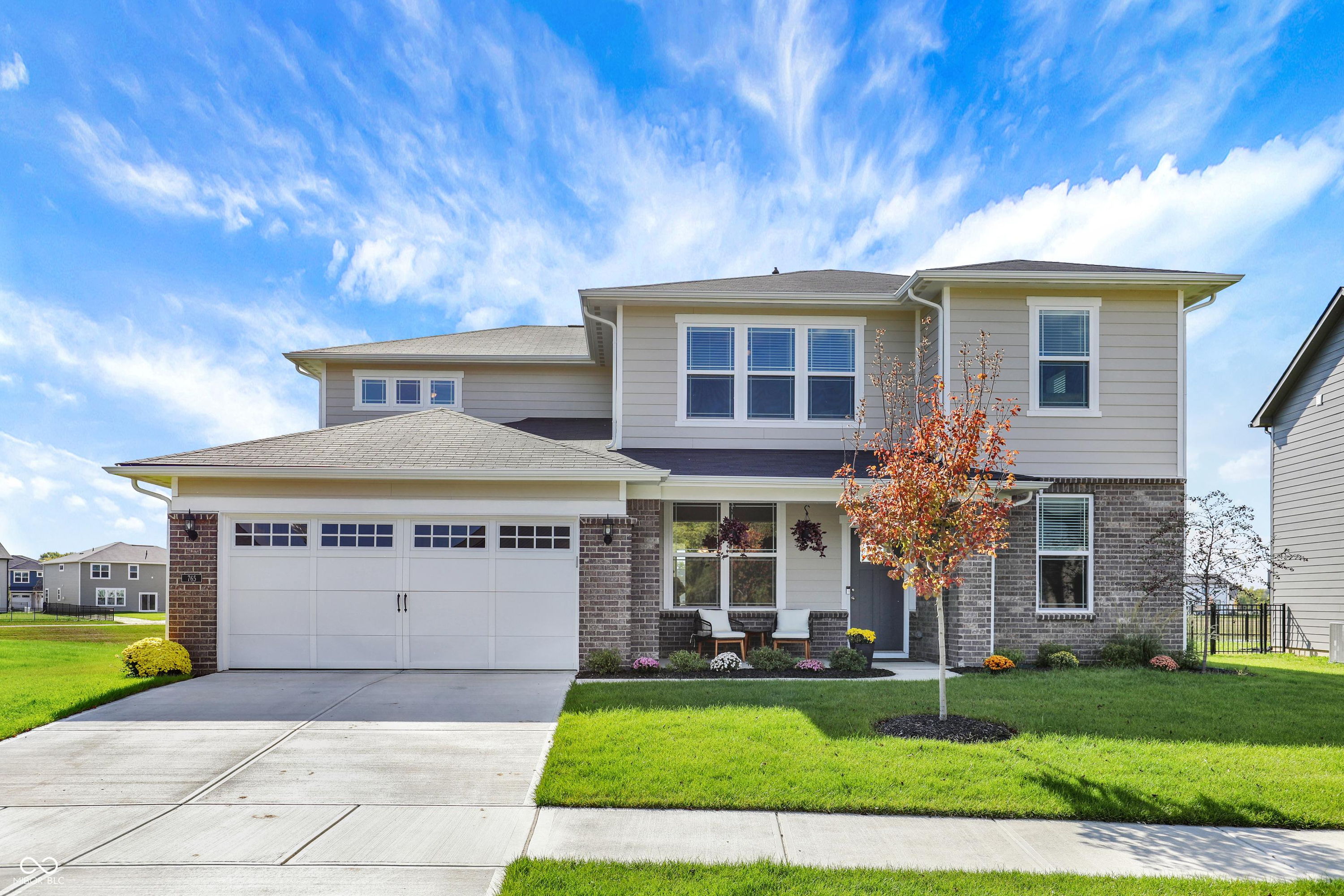Modern two-story house with stone siding, large windows, a porch with plants, and an autumn tree, under a bright sky.