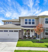 Modern two-story house with stone siding, large windows, a porch with plants, and an autumn tree, under a bright sky.