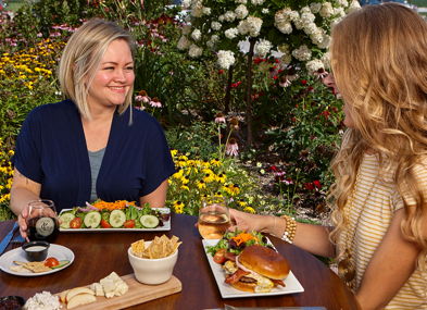 Two women enjoy a joyful outdoor meal in a vibrant garden, sharing sushi and burgers, smiling in each other's company.