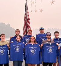 A smiling group in blue shirts stands by water, with a flag backdrop, exuding unity and celebration.