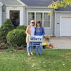 Two smiling women hold a "YARD MONTH" sign in front of a light blue house, celebrating autumn with greenery and decorations.
