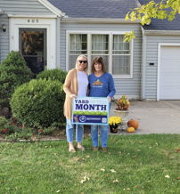 Two smiling women hold a "YARD MONTH" sign in front of a light blue house, celebrating autumn with greenery and decorations.