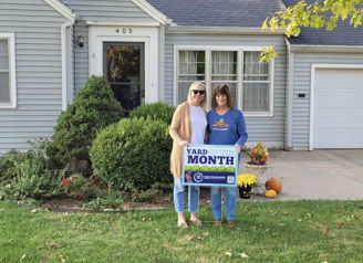 Two smiling women hold a "YARD MONTH" sign in front of a light blue house, celebrating autumn with greenery and decorations.