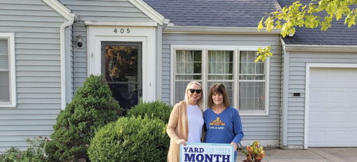 Two smiling women hold a "YARD MONTH" sign in front of a light blue house, celebrating autumn with greenery and decorations.
