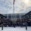 A united group in black shirts poses outside a building, radiating positivity and camaraderie under a partly cloudy sky.
