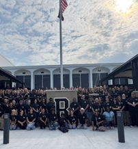 A united group in black shirts poses outside a building, radiating positivity and camaraderie under a partly cloudy sky.