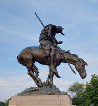 Bronze statue of a poised warrior on a dynamic horse, embodying strength and bravery against a tranquil sky backdrop.