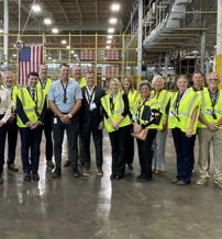Group in yellow safety vests in a warehouse, showing teamwork and positivity, with an American flag in the background.