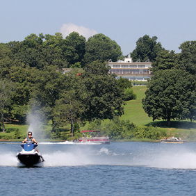 Two people ride jet skis on a serene lake, surrounded by green hills and a house, capturing adventure and summertime fun.