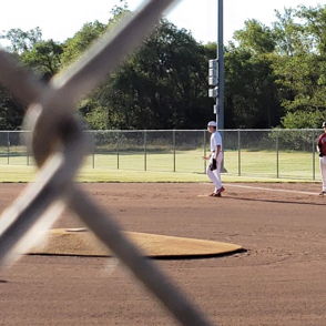 Baseball field scene through a chain-link fence, with players in uniform, evoking excitement, camaraderie, and nostalgia.