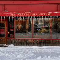 Cozy pizza restaurant with a red awning claims "BEST PIZZA IN THE UNIVERSE!" Icicles and snow create a chilly winter scene.