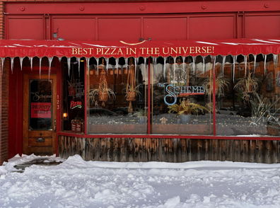 Cozy pizza restaurant with a red awning claims "BEST PIZZA IN THE UNIVERSE!" Icicles and snow create a chilly winter scene.