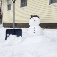 A cheerful snowman with a carrot nose and brown hat stands in a snowy scene, with a light yellow house and snow shovel nearby.