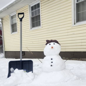 A cheerful snowman with a carrot nose and brown hat stands in a snowy scene, with a light yellow house and snow shovel nearby.