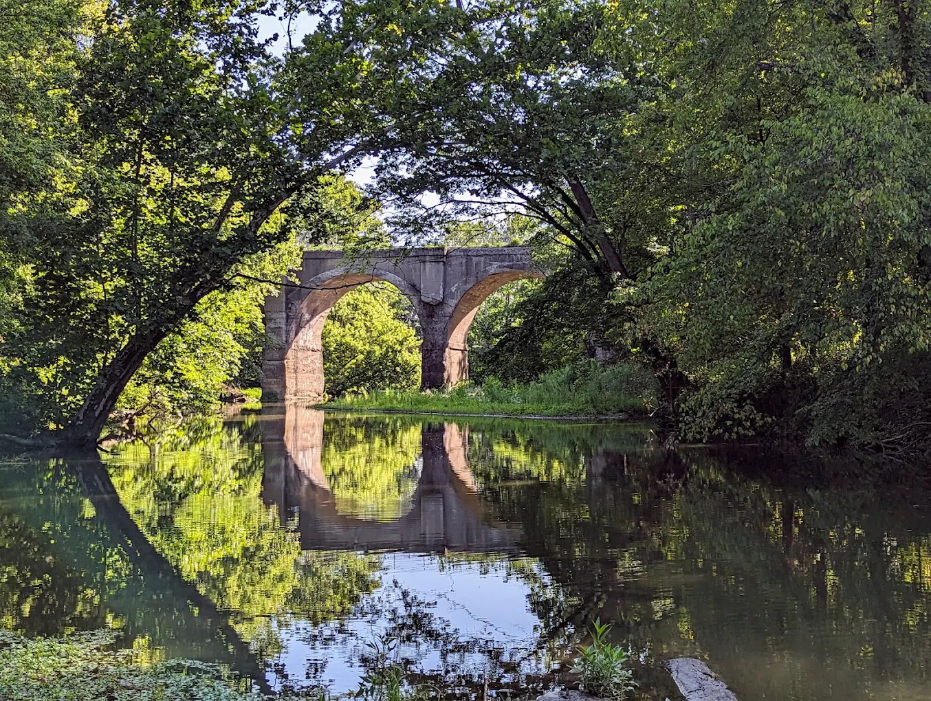 A serene landscape with a stone bridge over calm water, framed by lush greenery and reflections, evoking tranquility and nostalgia.