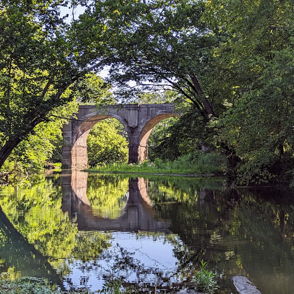 A serene landscape with a stone bridge over calm water, framed by lush greenery and reflections, evoking tranquility and nostalgia.