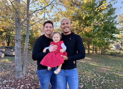 Two smiling adults stand outdoors with a child in a red dress, surrounded by autumn trees, evoking joy and togetherness.