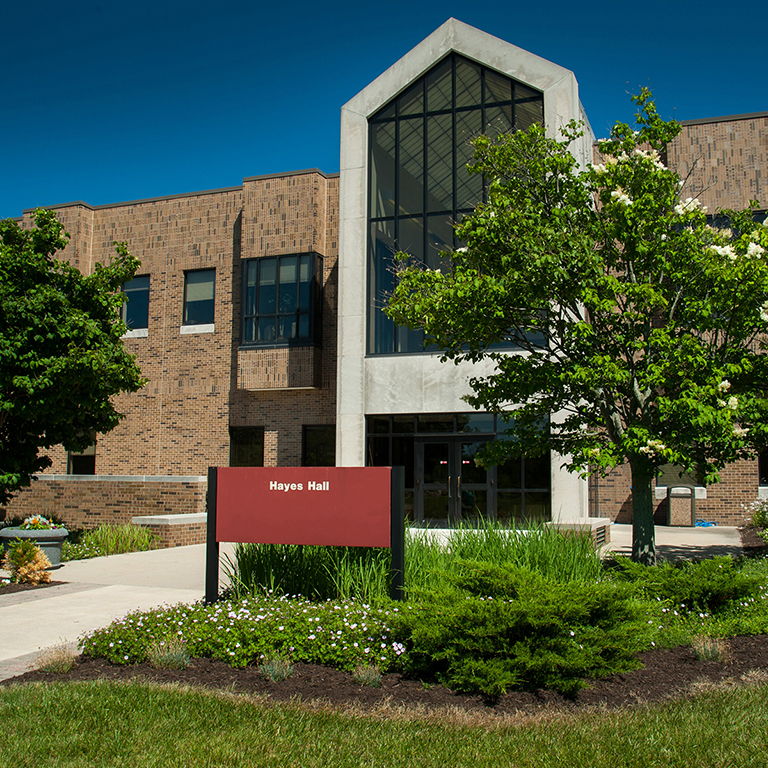 Modern building with glass entrance and "Hayes Hall" sign, surrounded by greenery, evoking positivity and tranquility.