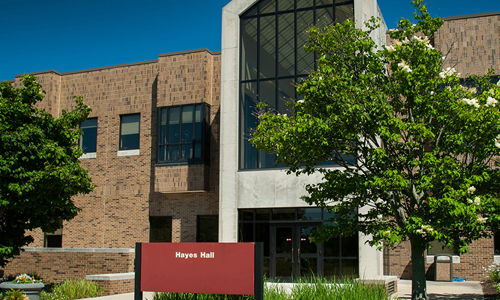 Modern building with glass entrance and "Hayes Hall" sign, surrounded by greenery, evoking positivity and tranquility.