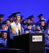 Graduation ceremony with a male speaker at a podium, graduates in blue gowns, evoking joy, excitement, and hope for the future.