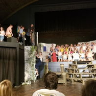 The image shows a lively theater performance with a large cast in dynamic poses, enhancing themes of excitement and collaboration.