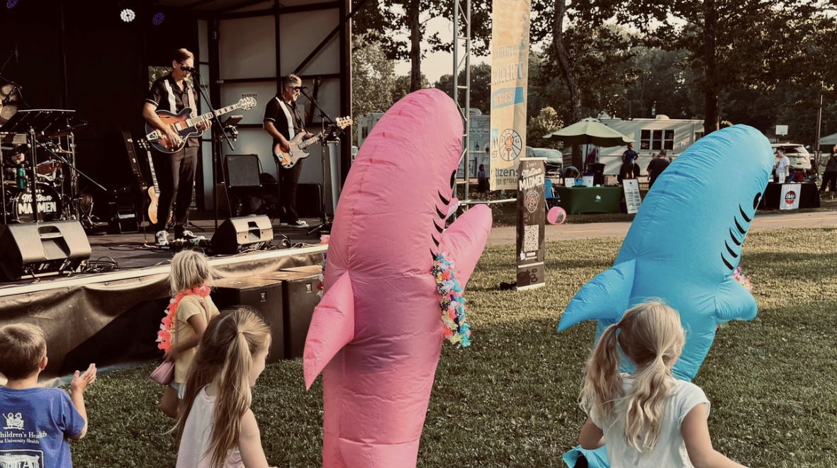 Children enjoy inflatable shark toys at a lively outdoor event as a band plays on stage, creating a festive atmosphere.